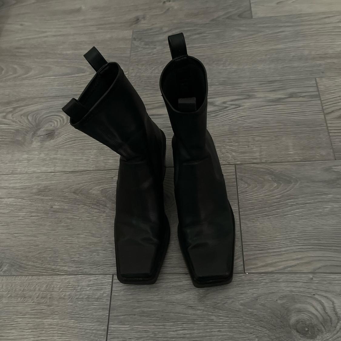 Black square-toed boots on grey wooden flooring, viewed from above.
