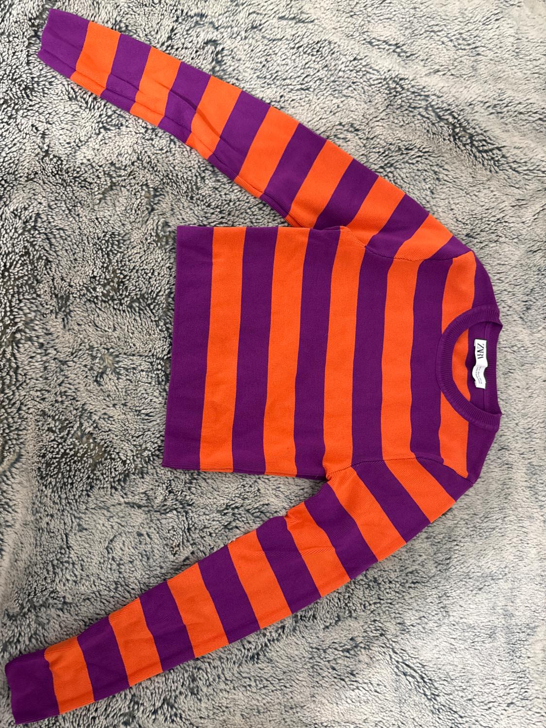 Purple and orange horizontal striped jumper on a grey textured surface. Visible label on the collar.