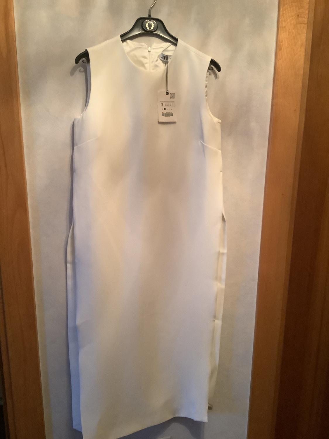 Sleeveless white dress hanging on a black hanger with a visible tag. Light wall background and wooden frame.