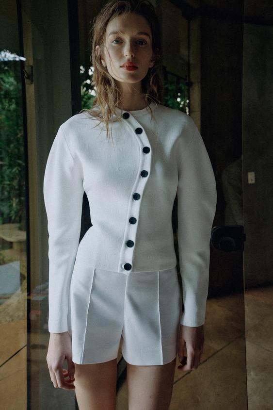 Sada bílého pleteného svetru s kulatým výstřihem, dlouhými rukávy a asymetrickým zapínáním na černé knoflíky, a bílých šortek.