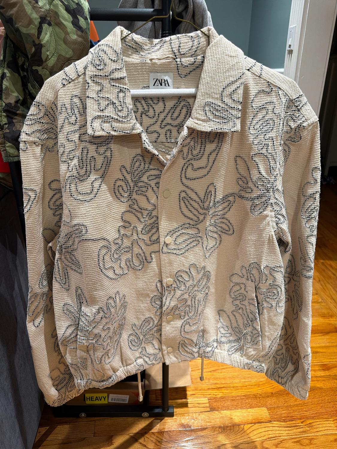 Beige Zara jacket with a shirt-style collar, button closure, and an abstract print of black lines, hanging on a hanger.