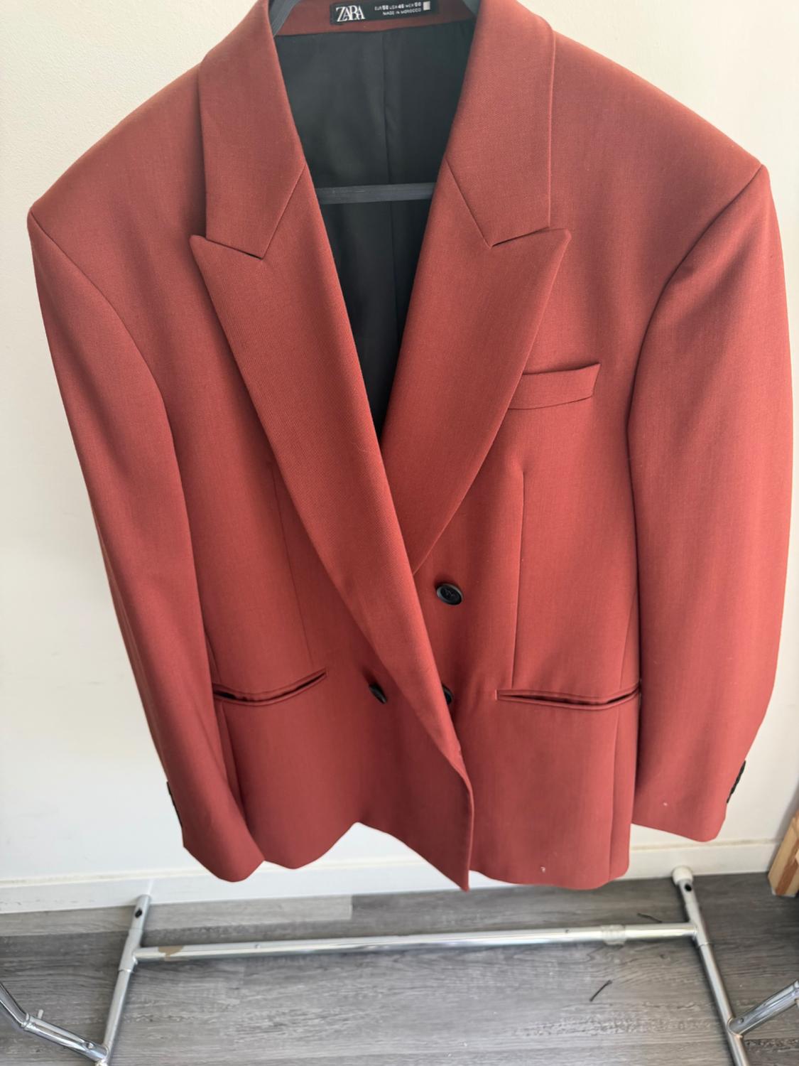 Red double-breasted jacket hanging on a hanger. Visible Zara label. White background and grey wooden floor.