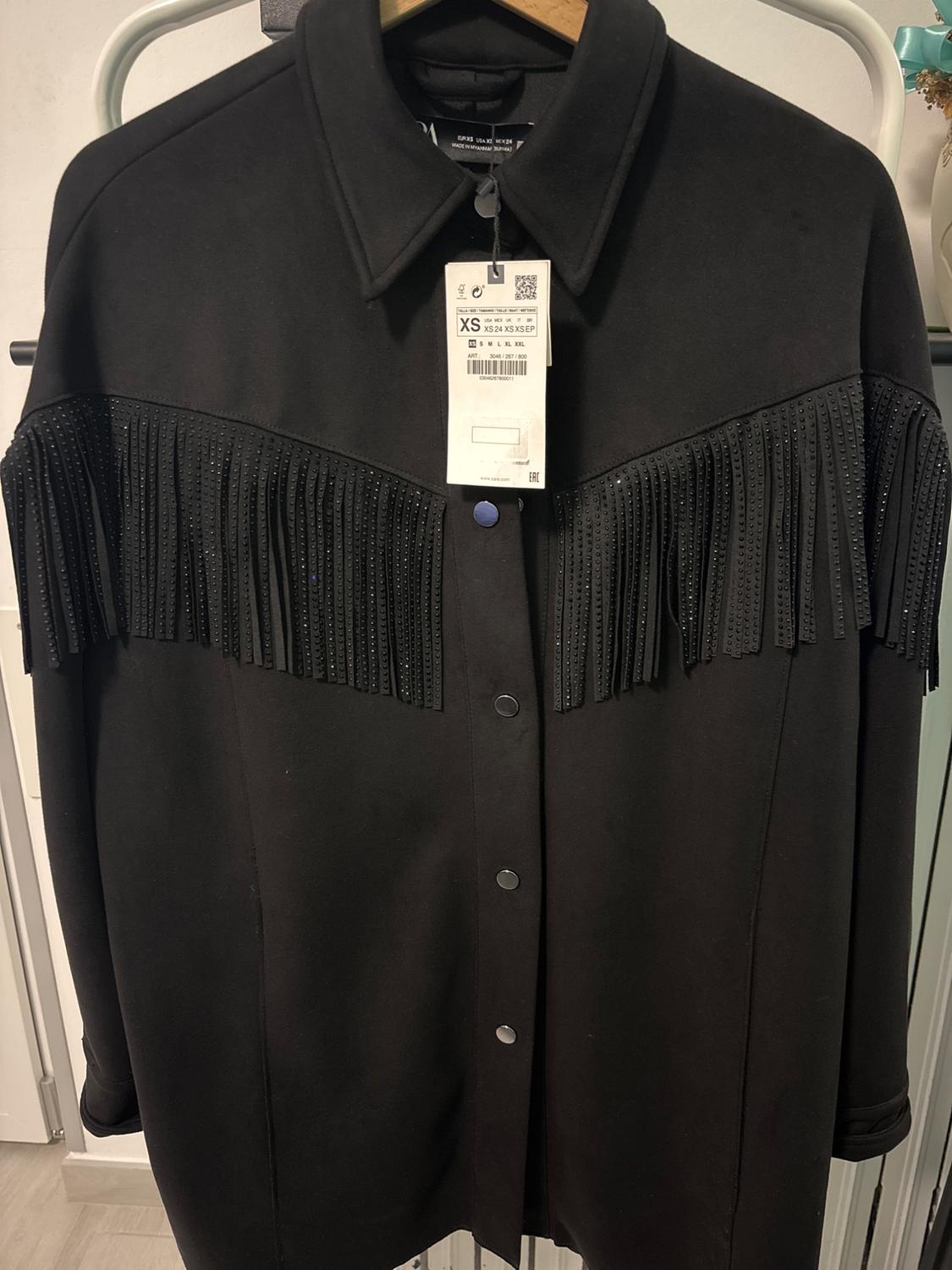 Black jacket with fringe and press studs, hanging on a hanger. Visible label with size XS and barcode.
