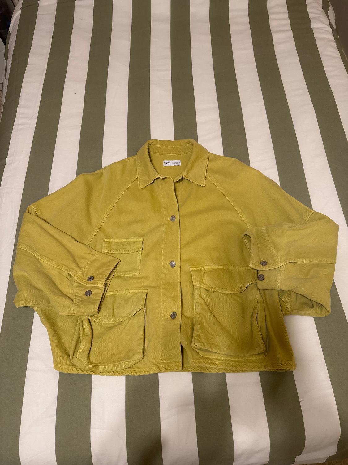 Yellow jacket with large pockets on a bed with a green and white striped bedspread. Label visible on the collar.