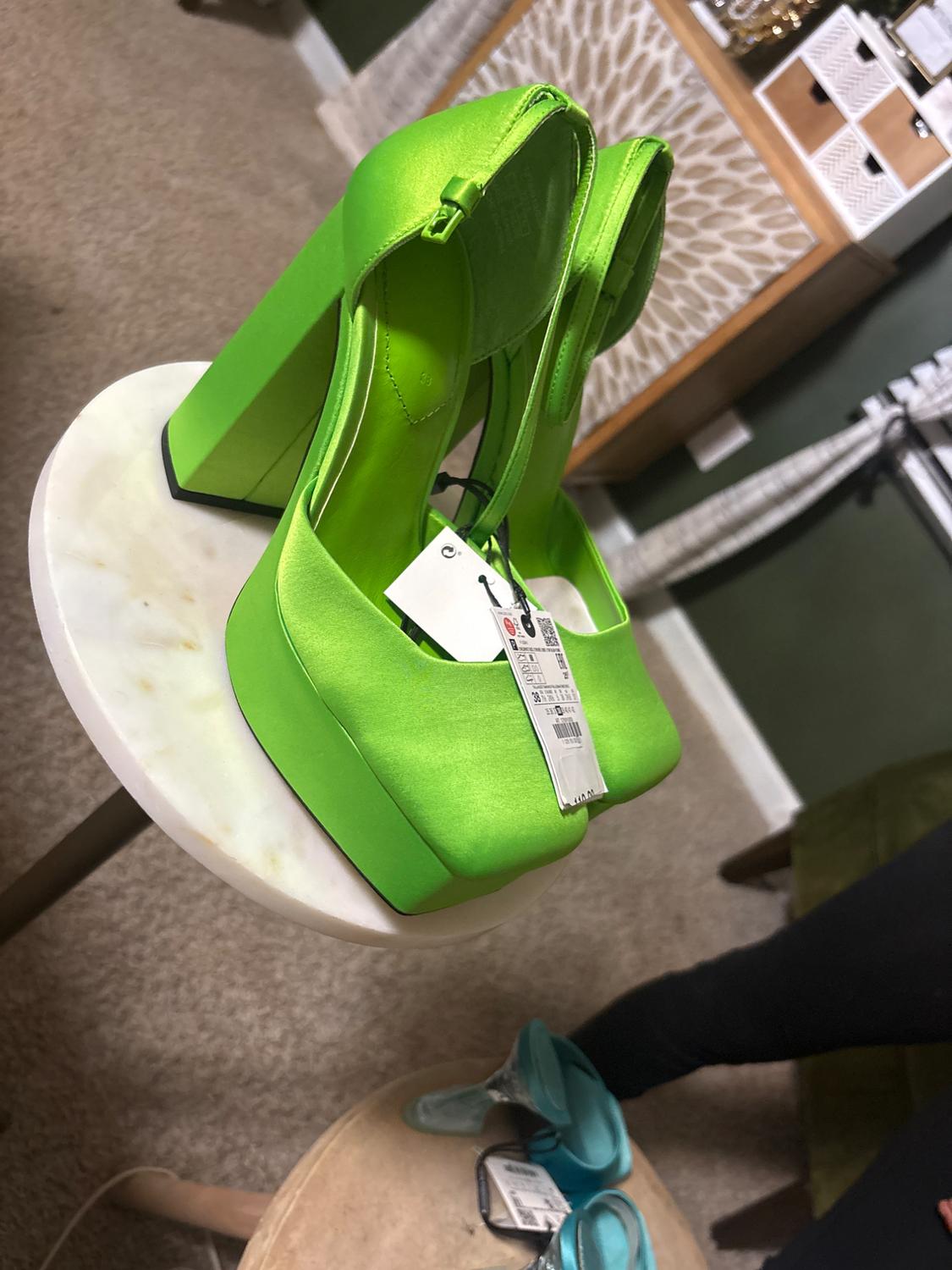 Neon green high heels on a white round table. Background with beige furniture and carpet. Visible tag on the shoes.