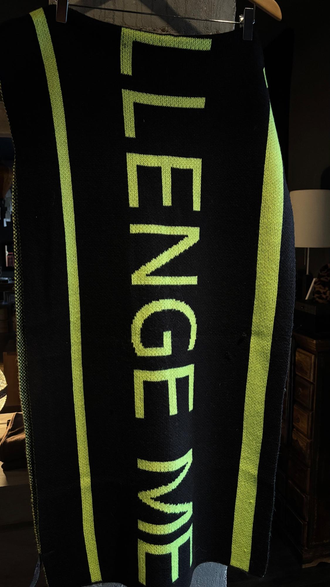 Black scarf with green stripes and vertical green text "CHALLENGE ME". Hung on a wooden hanger in an indoor setting.