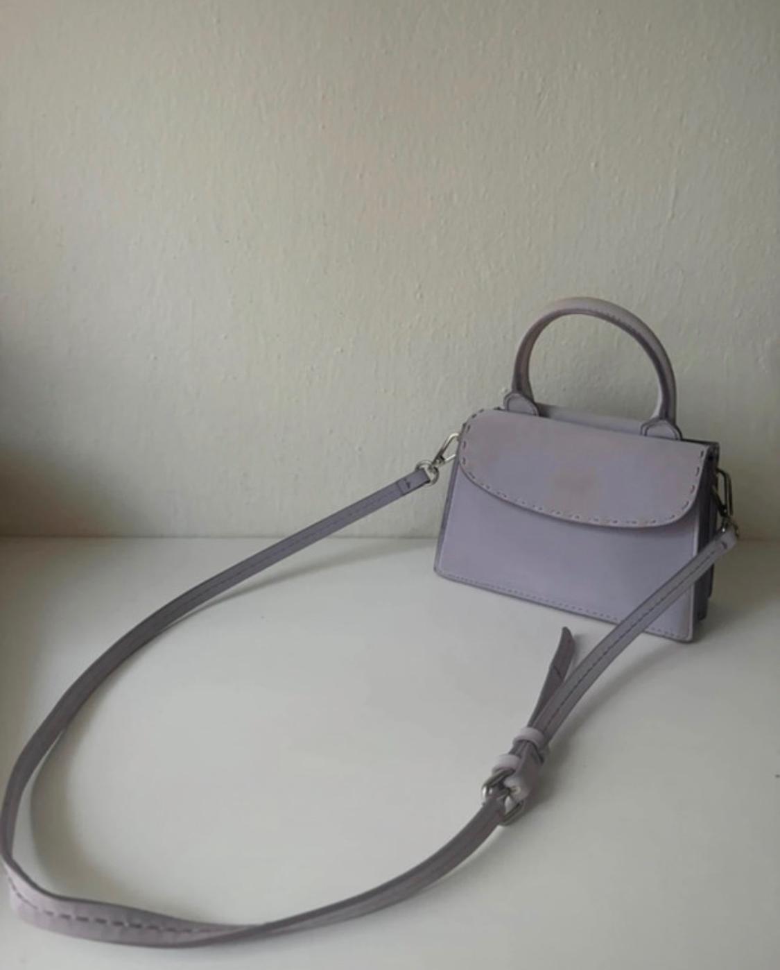 Small lilac handbag with a handle and long strap, on a white surface in front of a plain wall. Minimalist composition.