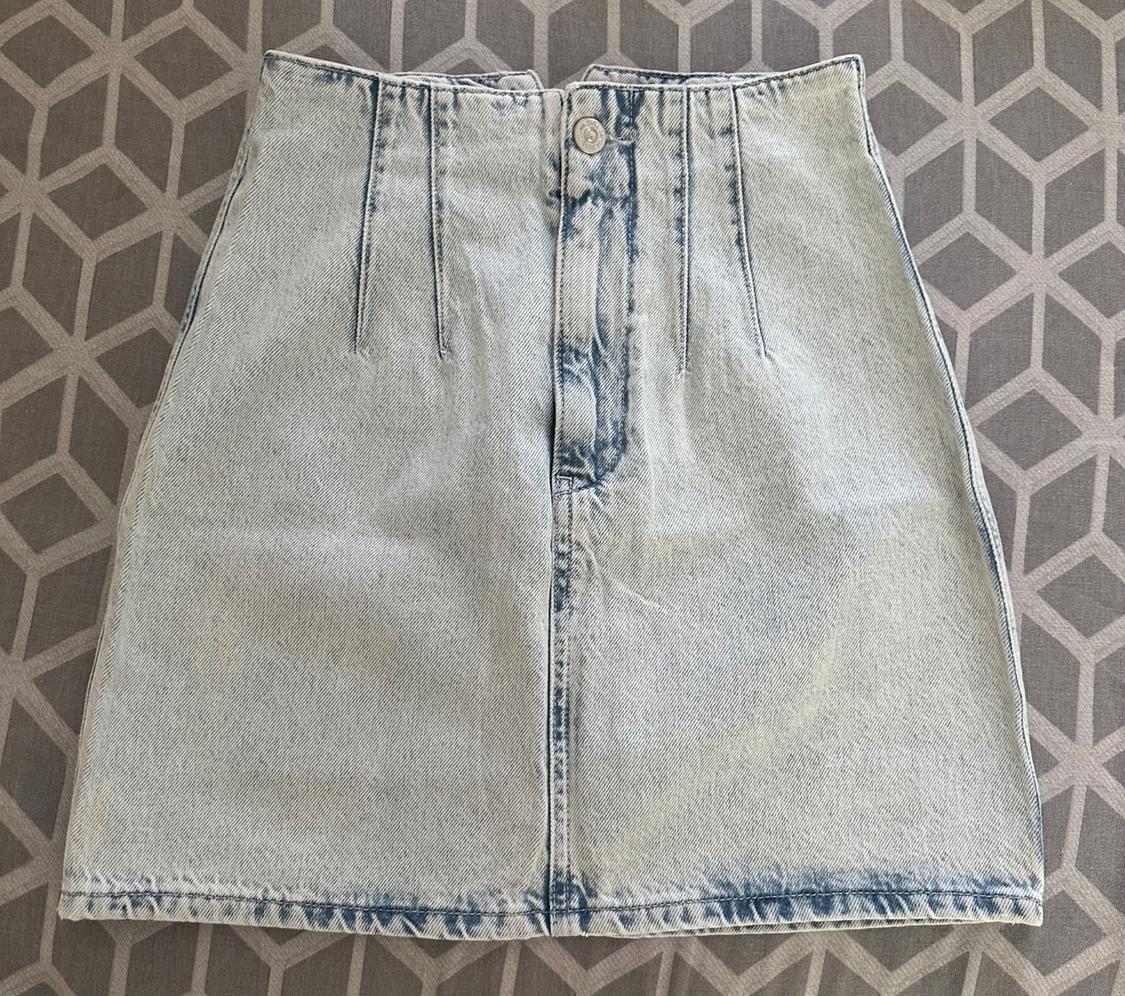 Light blue high-waisted denim miniskirt with front seams, set against a grey patterned background.