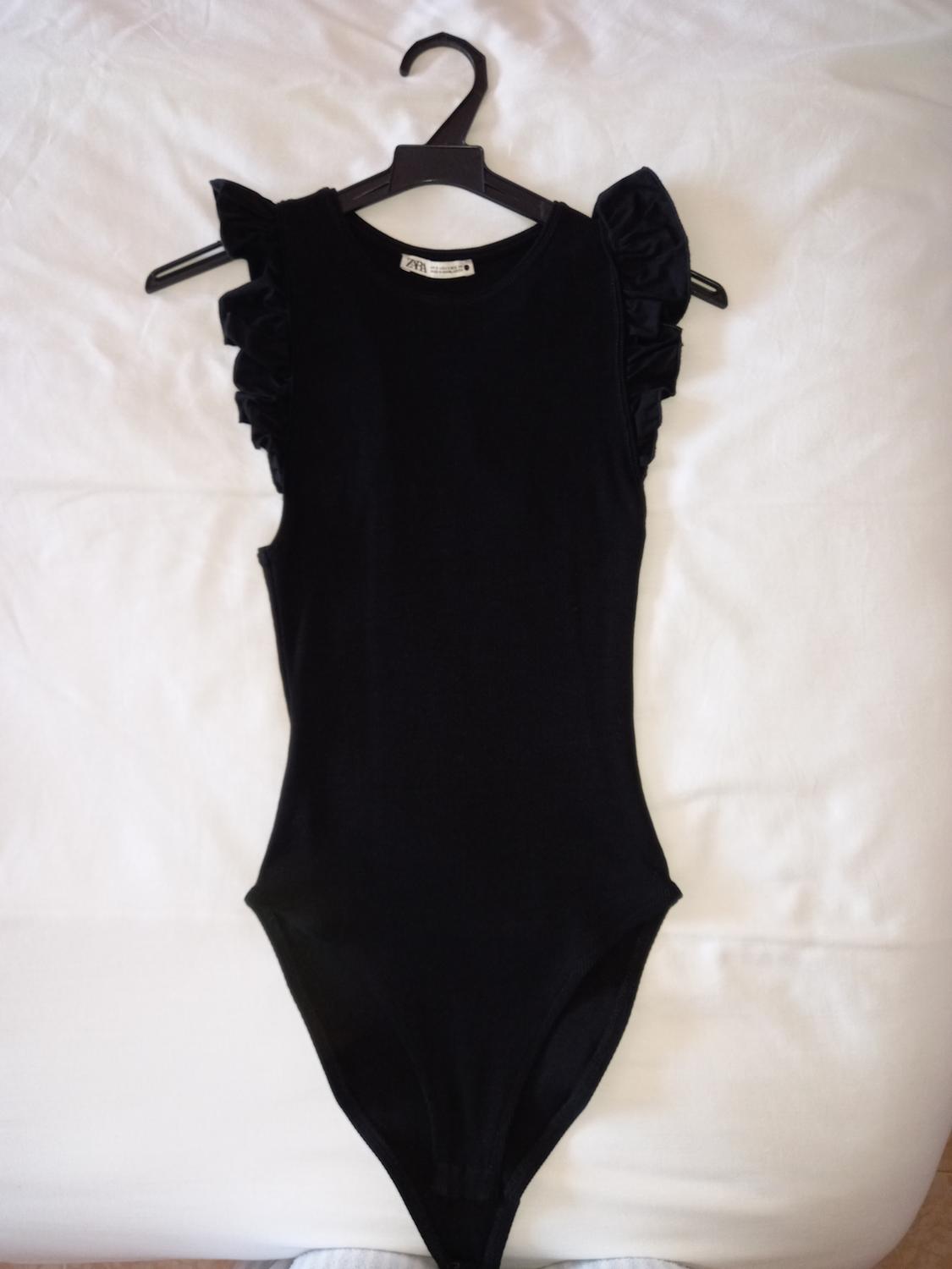 Sleeveless black bodysuit with frill shoulders, hanging on a hanger against a white background.