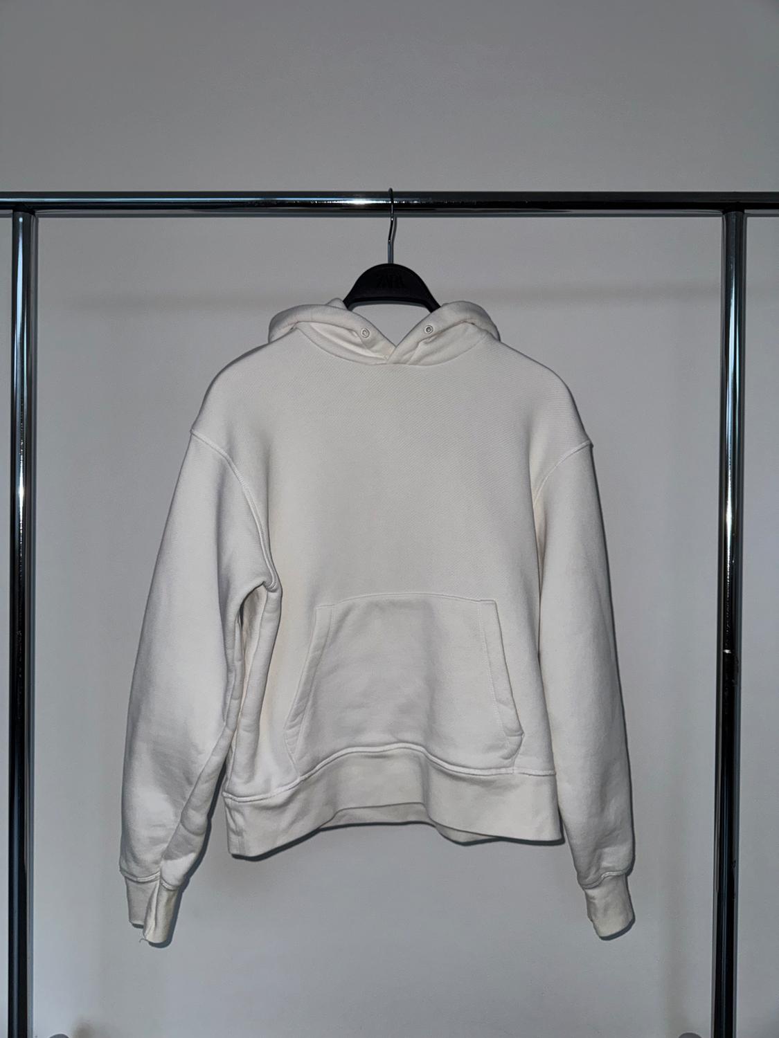 White hooded sweatshirt hanging on a black hanger on a metal stand, viewed from behind against a light grey background.
