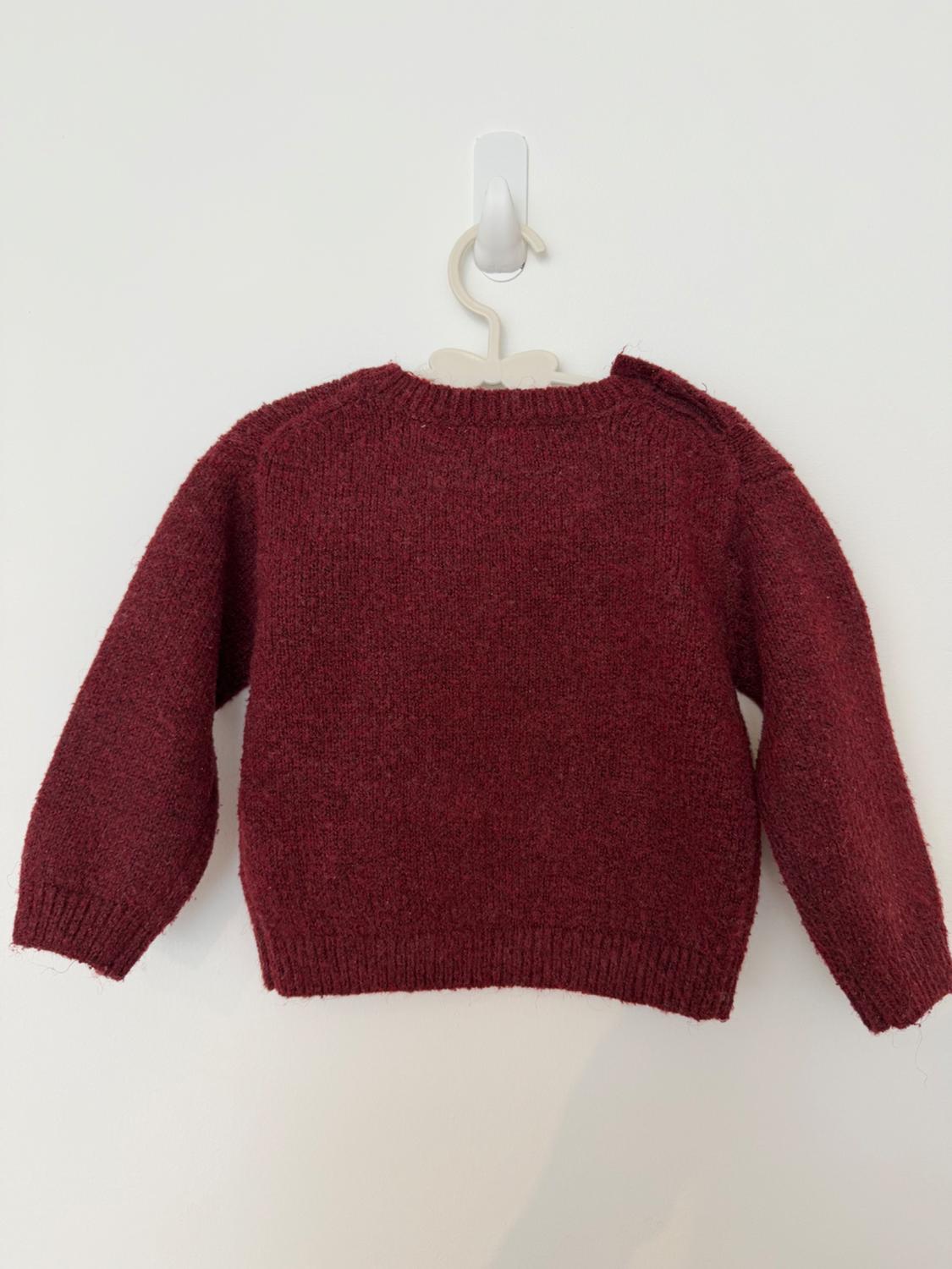 Maroon knitted jumper hanging on a white hanger against a plain wall. Simple composition with a relaxed style.