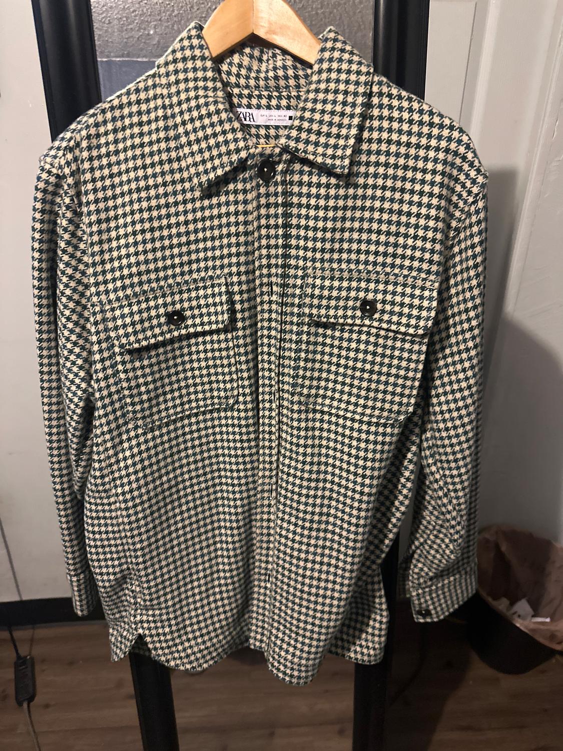 Black and white checked shirt hanging on a wooden hanger. Visible brand tag. White wall background and wooden floor.