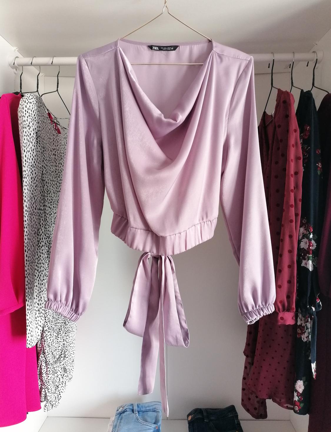 Lilac blouse with a draped neckline and a tie at the waist, hanging in a wardrobe next to other varied coloured garments.
