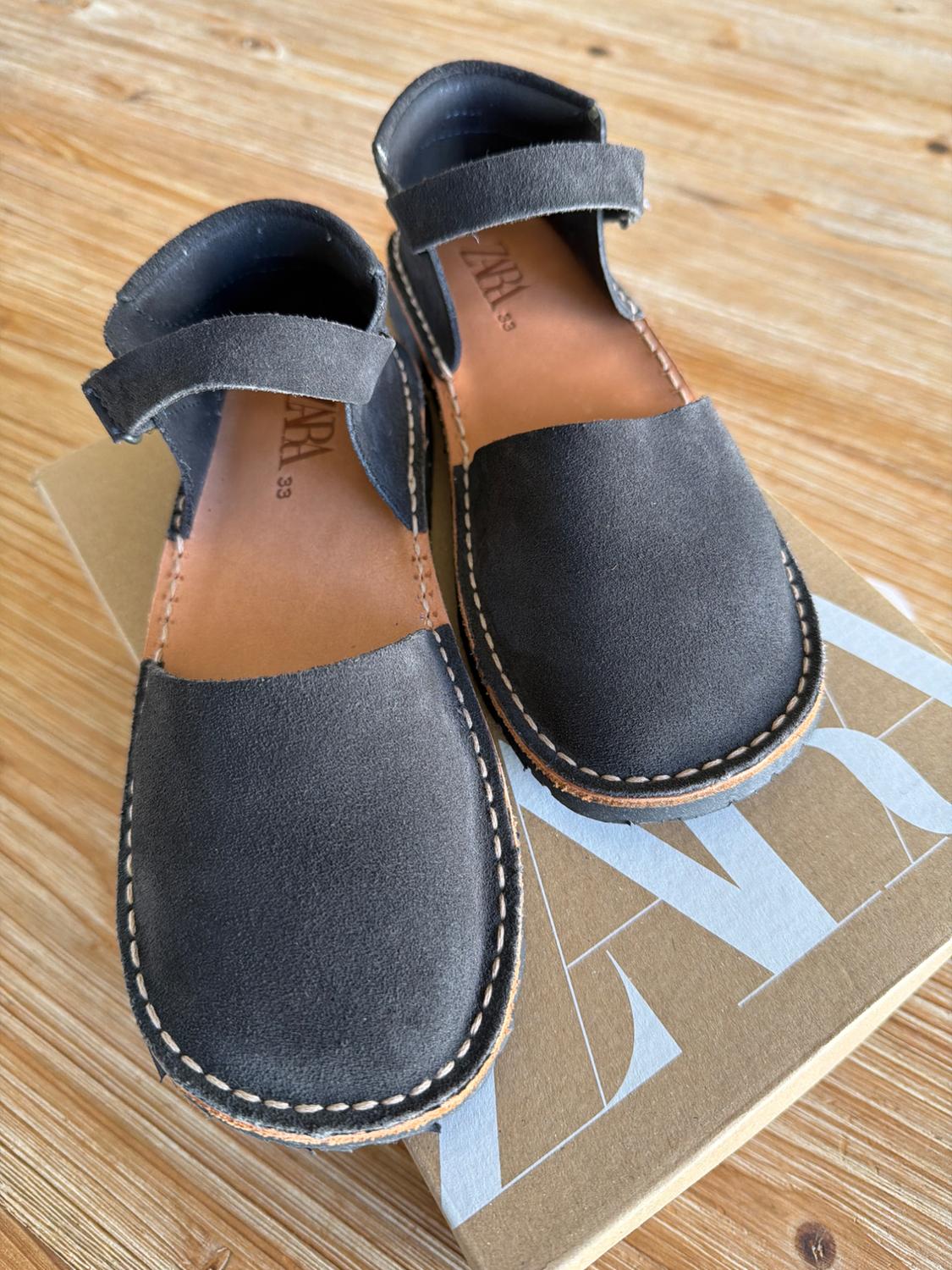 Black leather sandals with visible stitching on a cardboard box with the text "ZARA" on a wooden table.