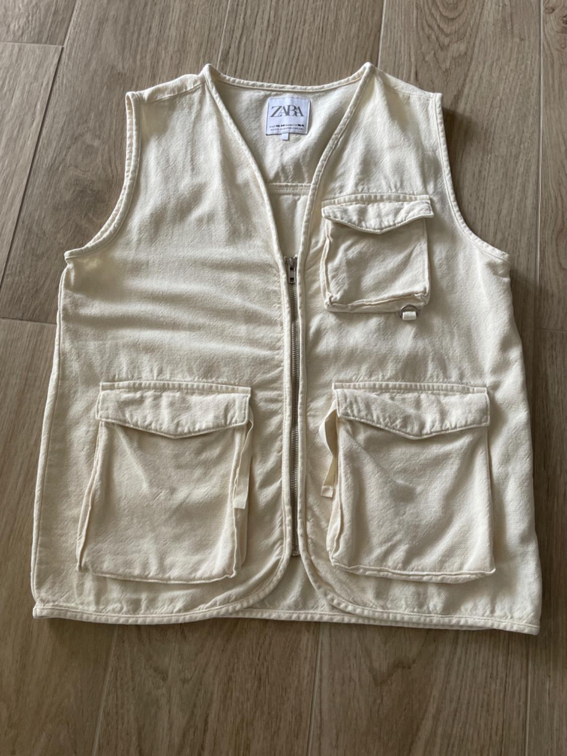 Beige sleeveless gilet with zip fastening and three large pockets on the front, on a wooden floor. Visible label: ZARA.