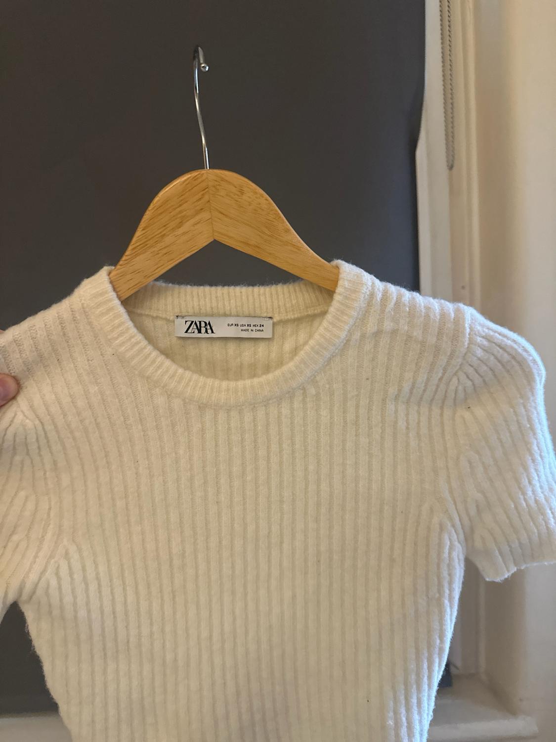 White ribbed short-sleeved knitted top on a wooden hanger. Label with the text ZARA EUR XS USA XS MEX 24 MADE IN CHINA.