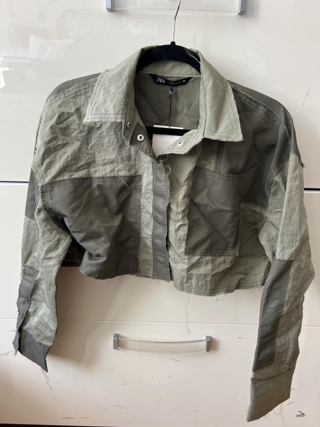 Olive green short jacket hanging on a black hanger in front of a white background. The tag shows the brand "Zara".