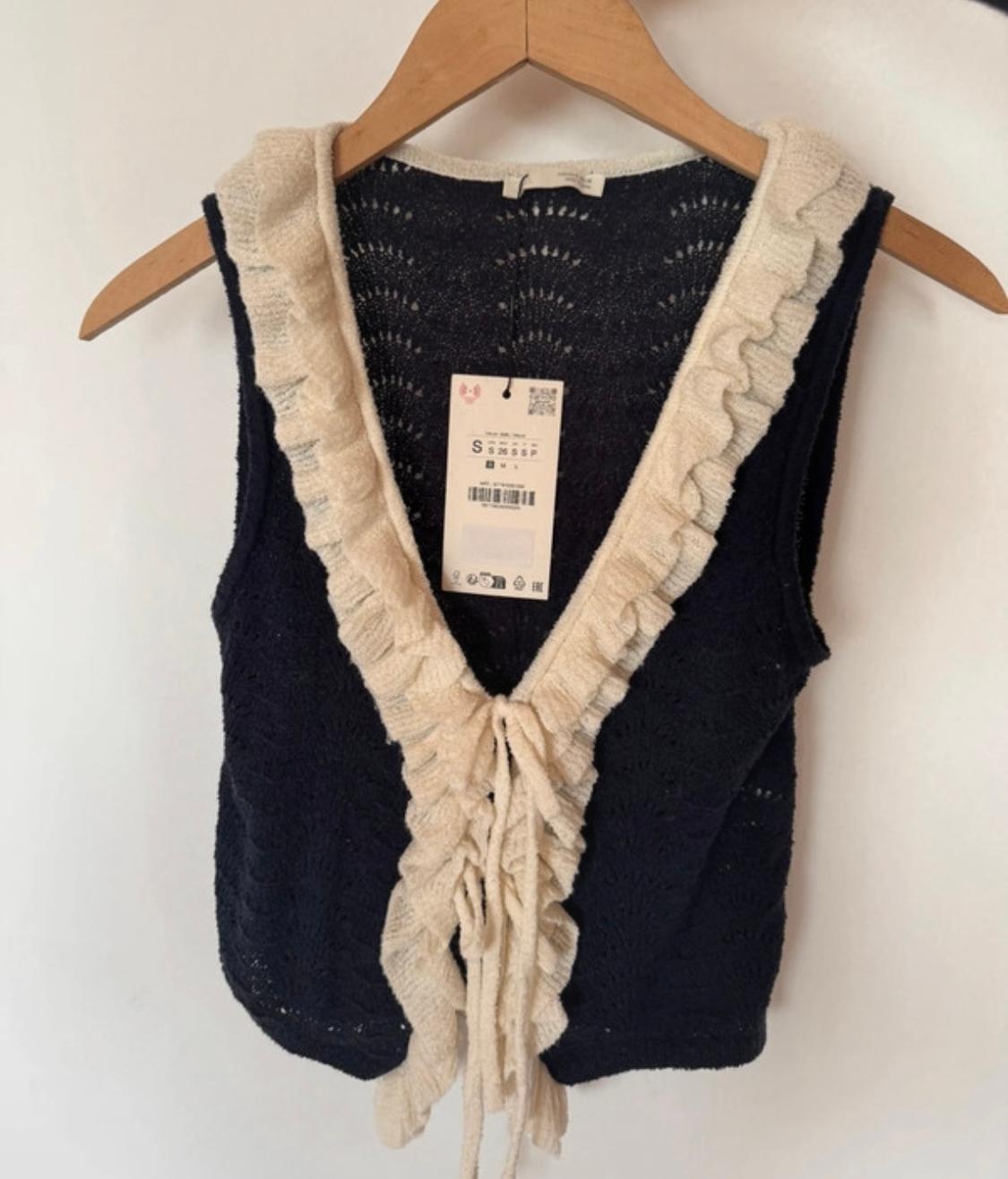 Black knitted waistcoat with beige ruffles on the trim, hanging on a wooden hanger. Visible label in the centre.
