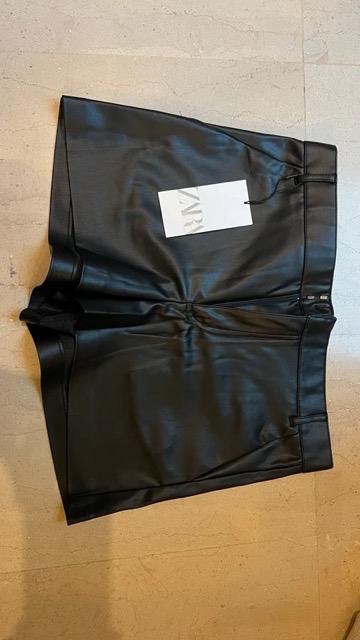 Black shorts made of shiny material on a light floor, with a white label that says "ZARA" placed on top.