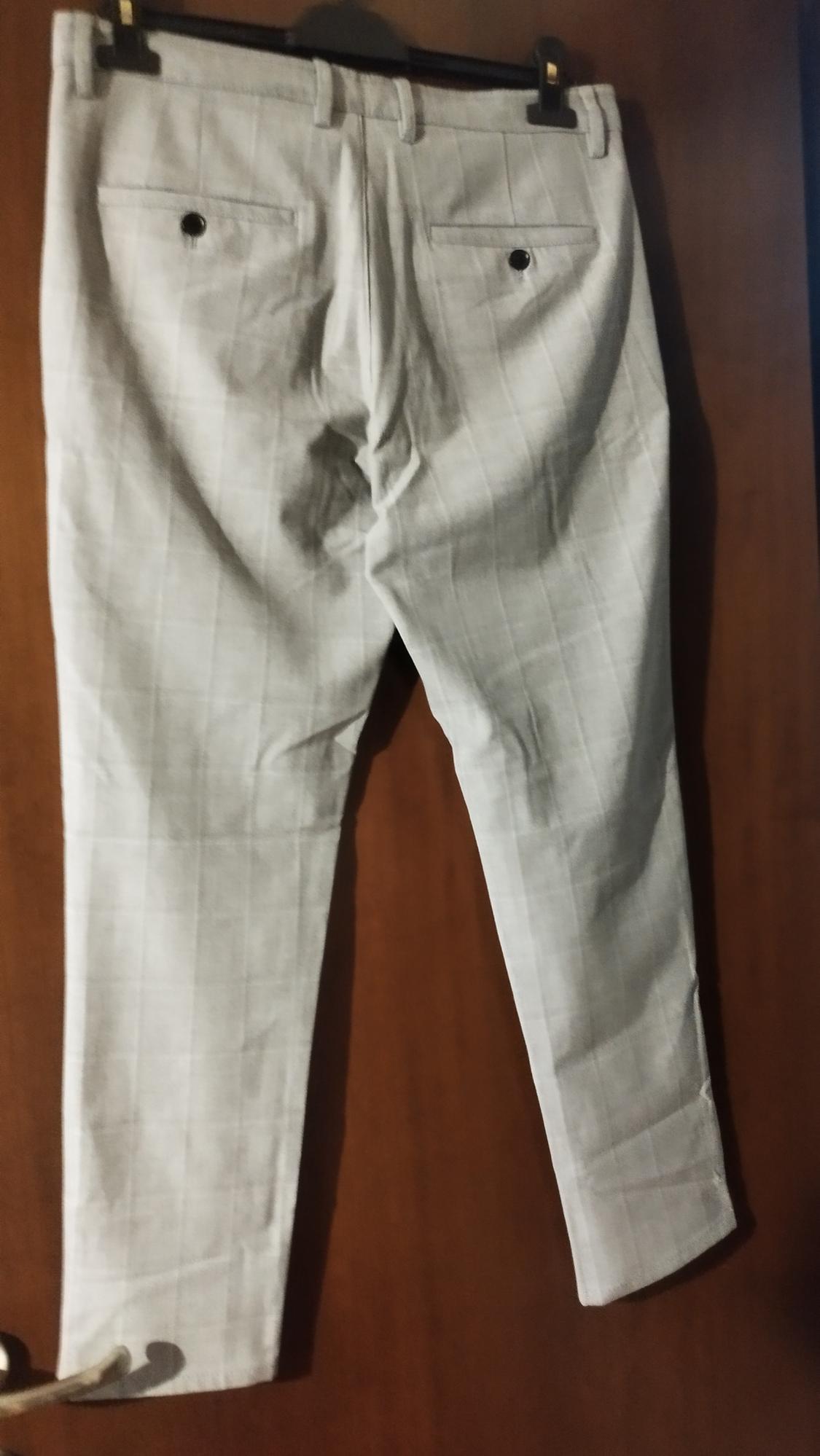 Light-coloured trousers with a check pattern hanging on a hanger over a wooden door. Rear pockets with buttons.