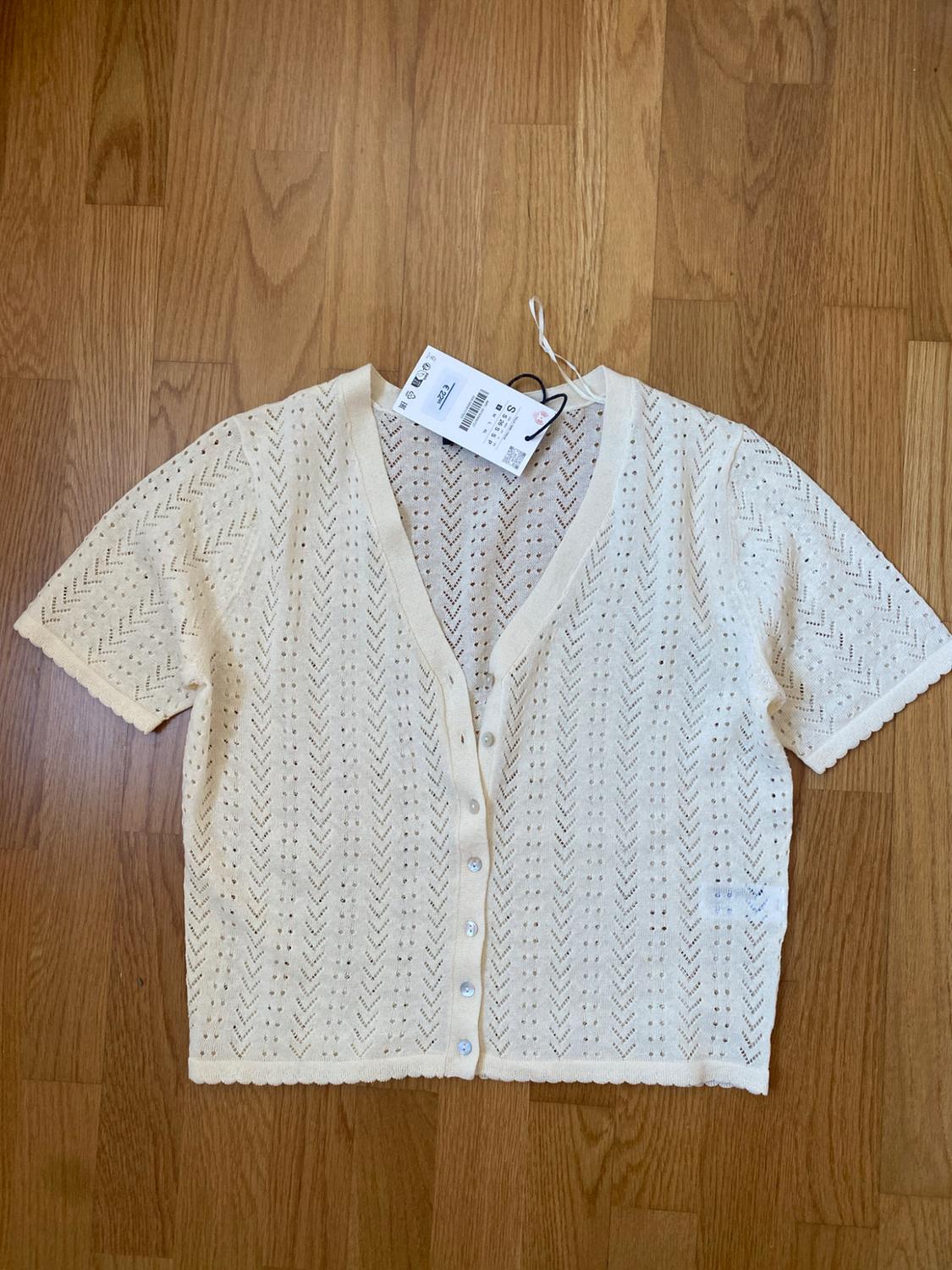 Cream open-knit cardigan, short sleeves, V-neck and buttons. Garment with tags on a wooden floor.