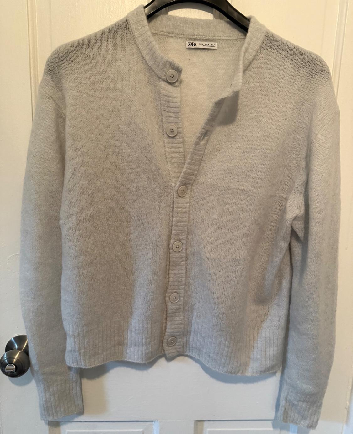 Light grey knitted cardigan hanging on a black hanger over a white door. Visible label with text "Zara".