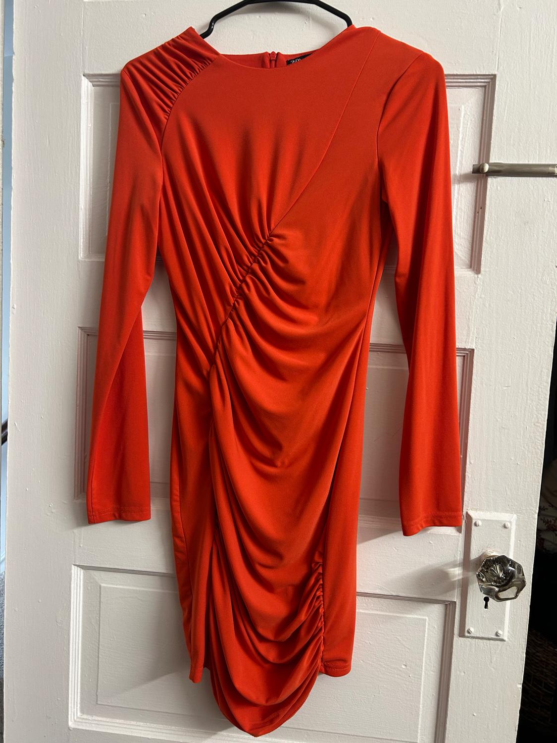 Long-sleeved orange dress with gathers at the front, hanging on a hanger over a classic white door with a glass knob.