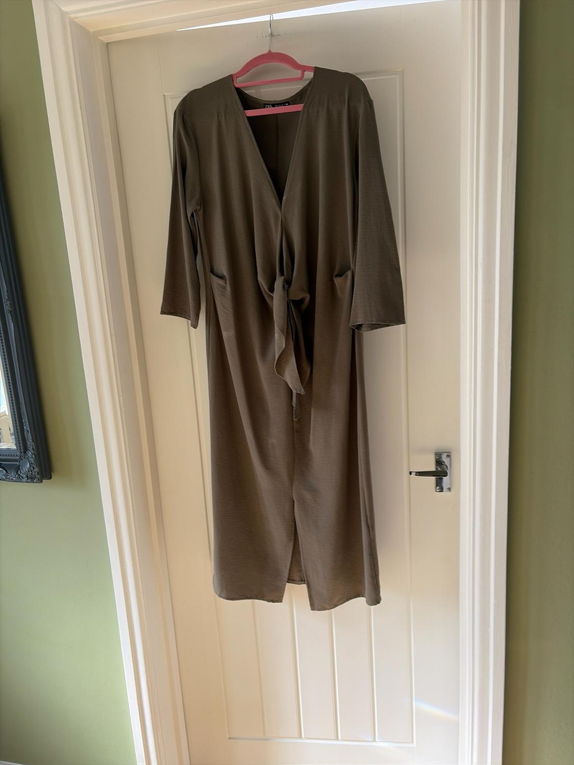 Olive green maxi dress with a V-neck, three-quarter sleeves and
front knot detail, hanging on a pink hanger on a white door.