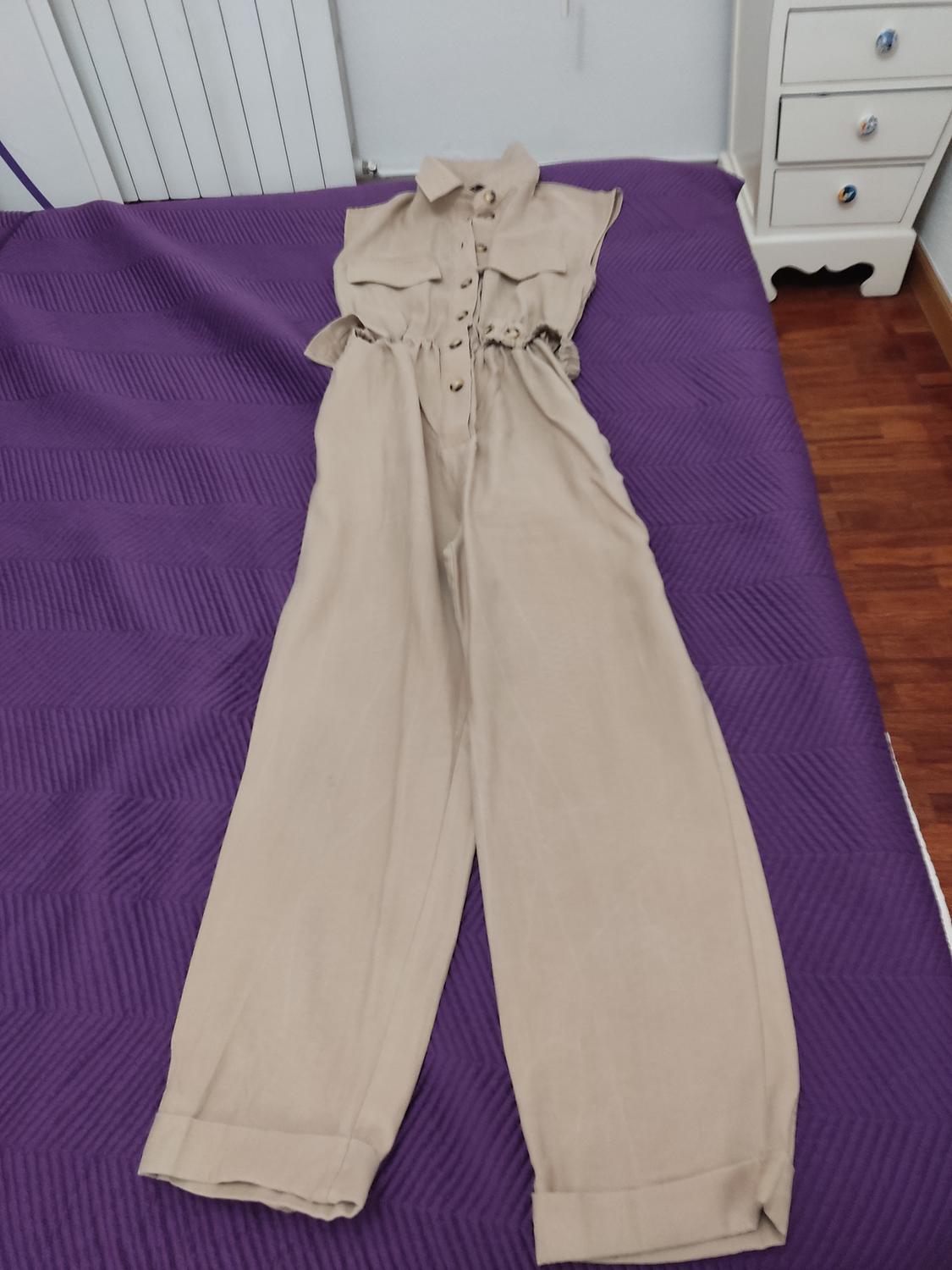Beige sleeveless jumpsuit with buttons down the front, on a bed with a purple bedspread. Background with white wall and white furniture.