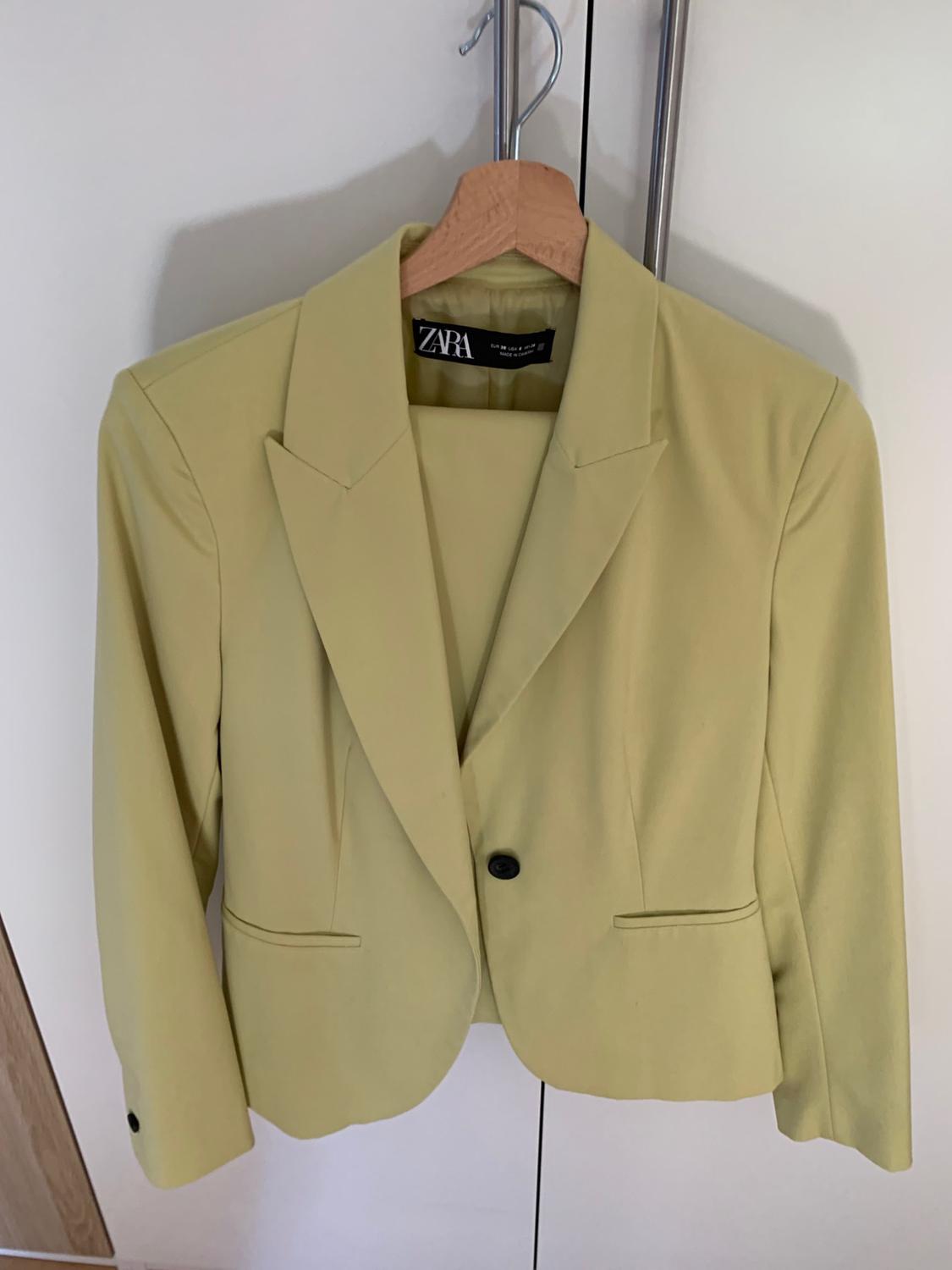 Light green suit consisting of a buttoned blazer and trousers, hanging on a wooden hanger in a white wardrobe. ZARA label visible.