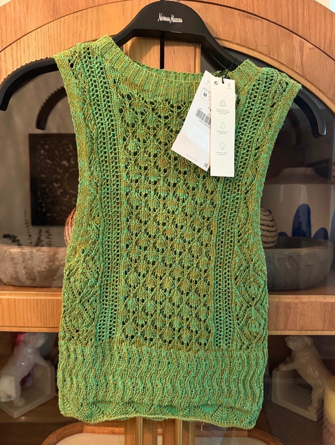 Green and yellow openwork top with label, hanging on a hanger bearing the text Neiman Marcus, in front of a wooden piece of furniture.