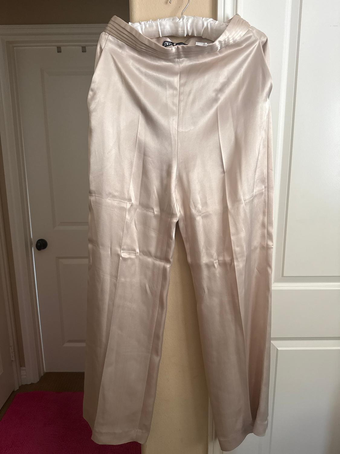 Cream-colored satin pants hanging on a hanger, with front pleats, in an indoor setting with white doors.
