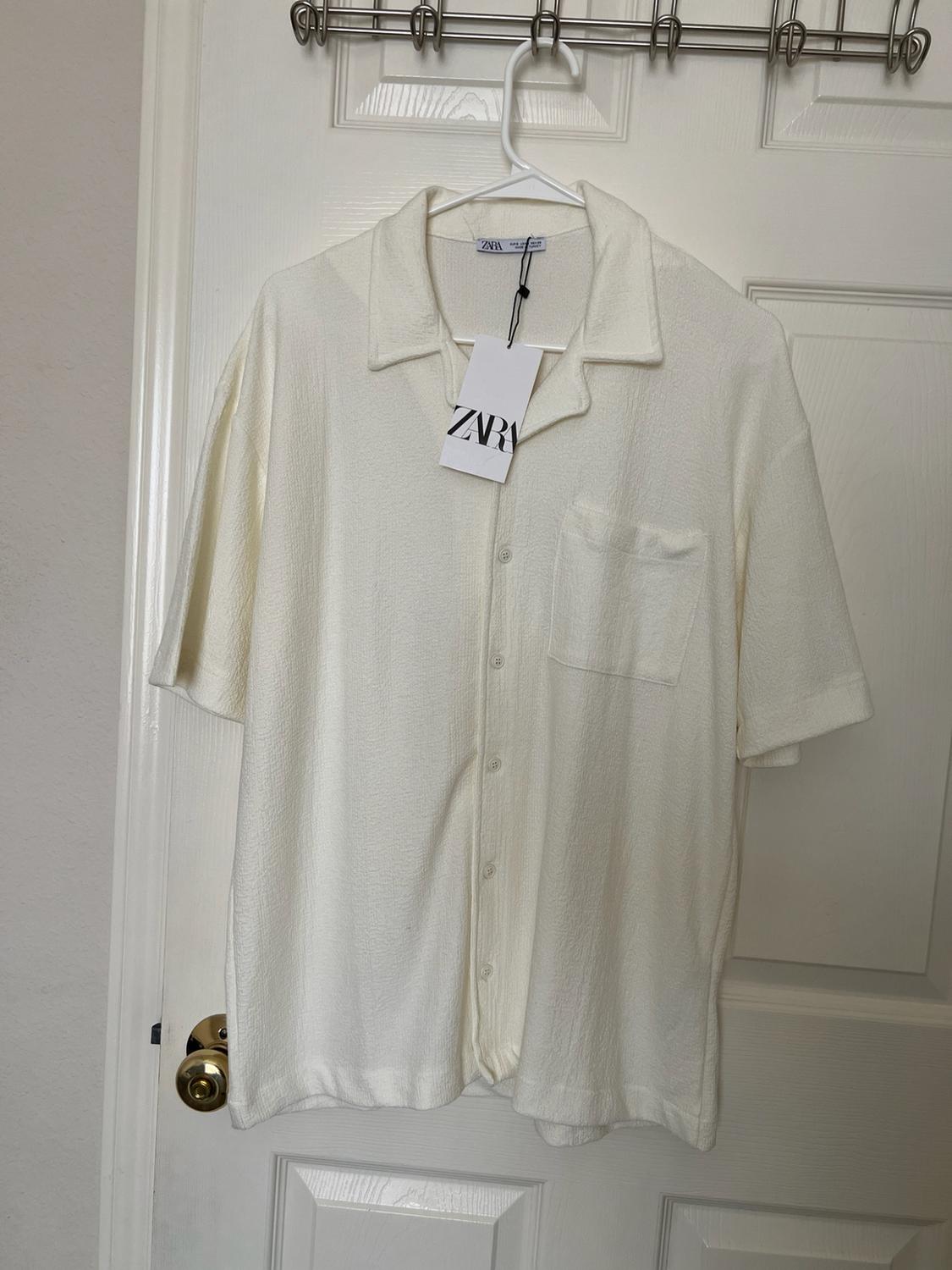 White textured short-sleeve shirt with a pocket, hanging on a door. Label with the text Zara.