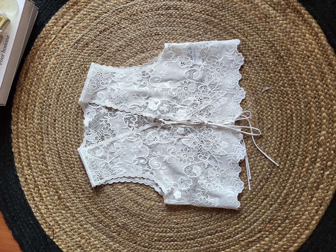 White lace corset with ties, on a round woven rug. To the left, a book and a perfume bottle.