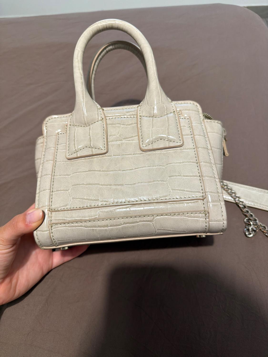 Hand holding a beige structured bag with a crocodile texture, short handles and a chain, on a brown fabric surface.