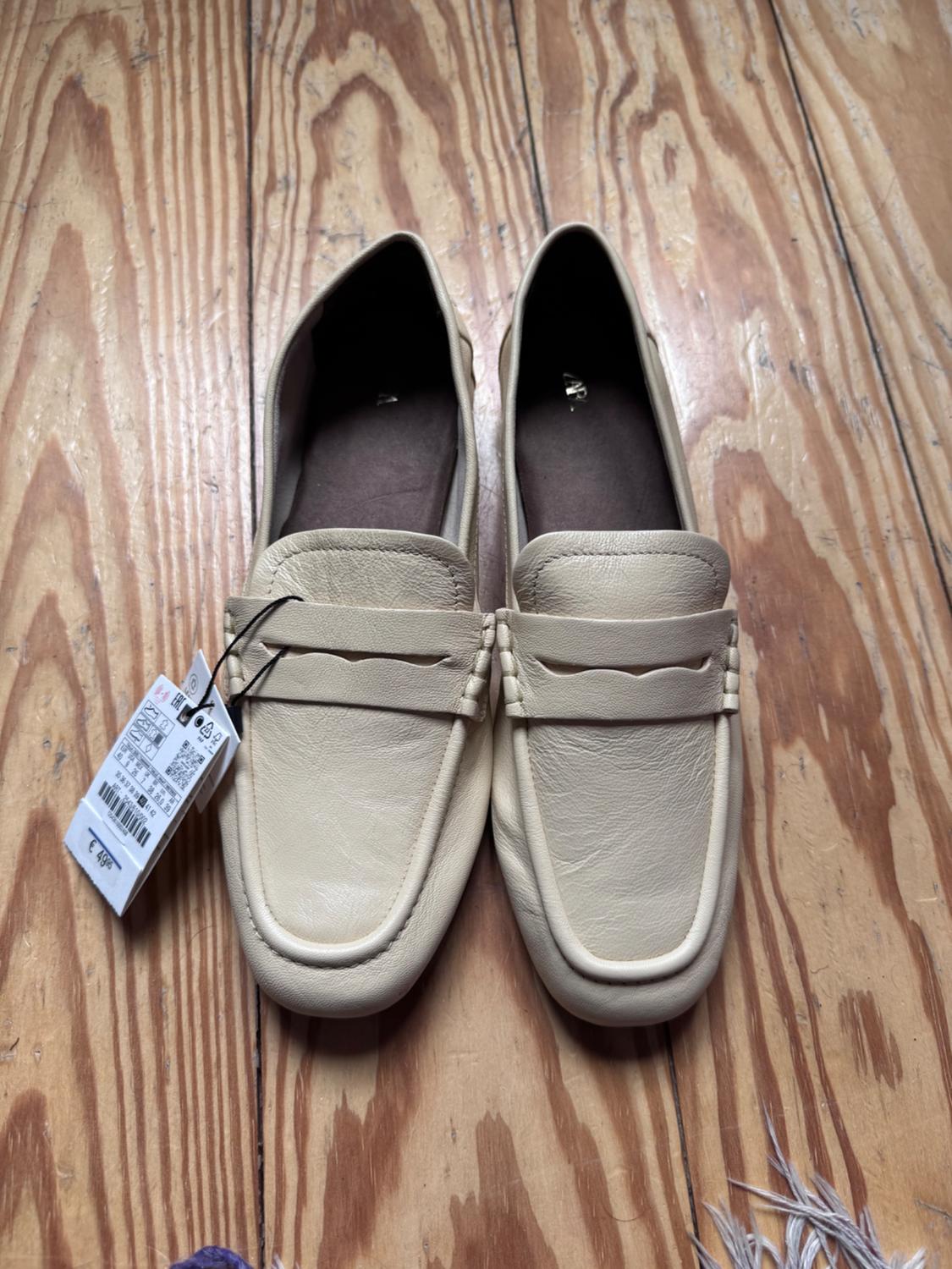 Top-down view of casual beige loafers on a wooden floor. The left shoe has a tag with the price €49.95.