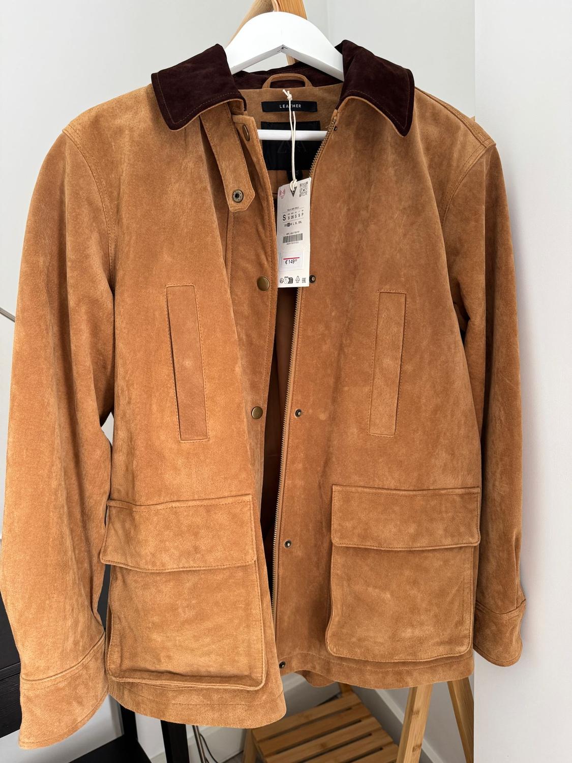 Light brown suede jacket with a dark collar, hanging on a white hanger. Visible label with product details.