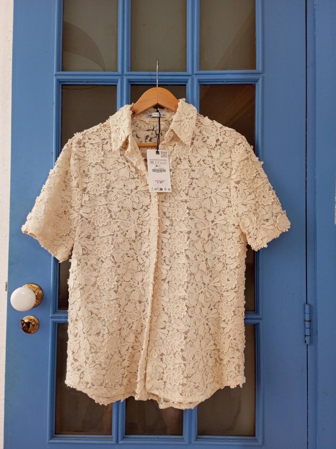 Cream floral lace shirt on a wooden hanger against a blue door with glass panels. Text: ZARA XS €39.