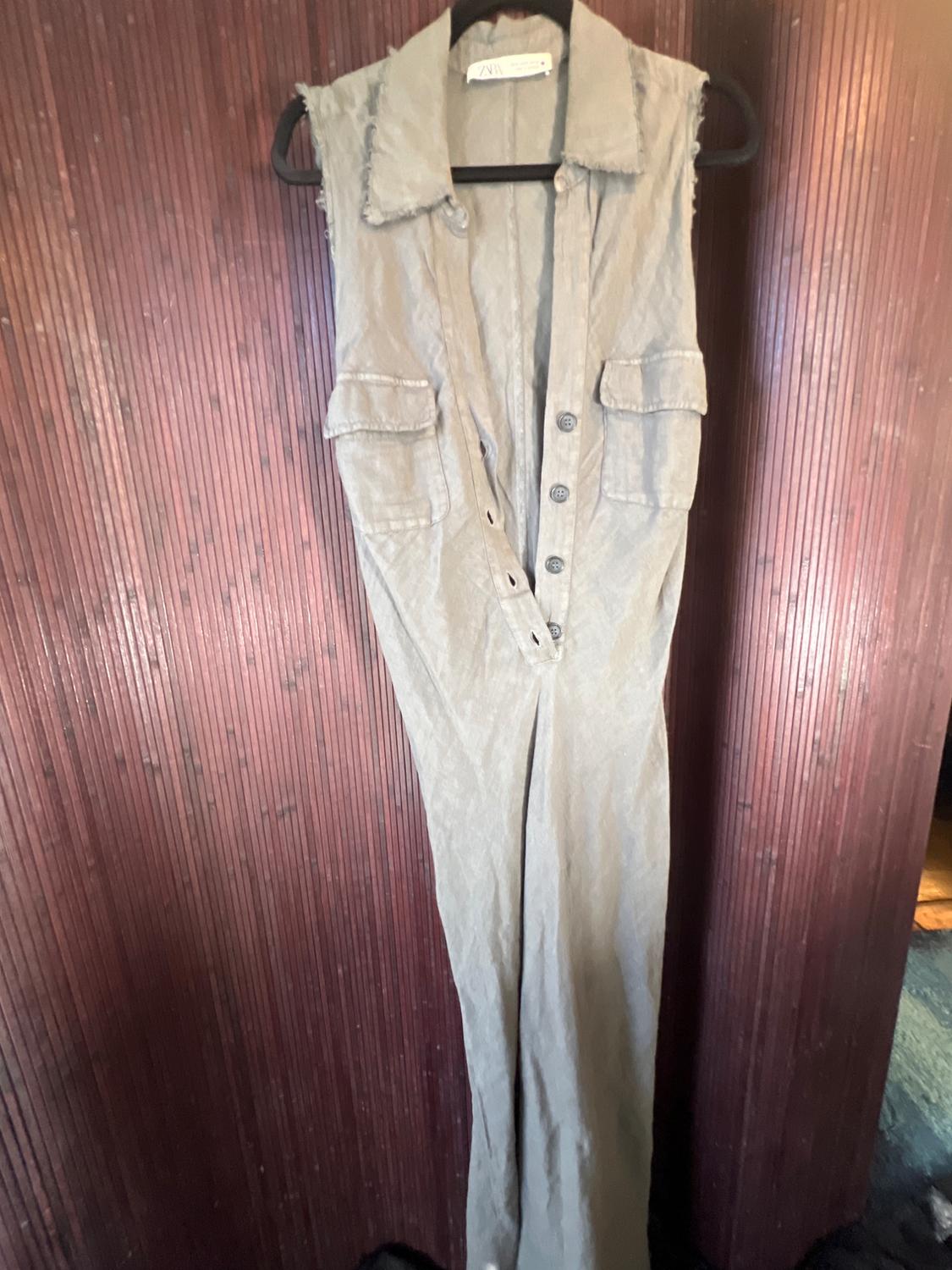 Beige sleeveless jumpsuit hanging on a black hanger against a brown wooden background. It has front pockets and buttons.