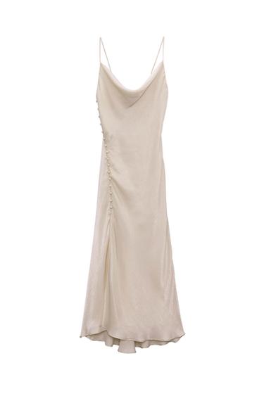 Long cream-colored spaghetti strap dress, with a draped neckline and decorative buttons on one side. White background.