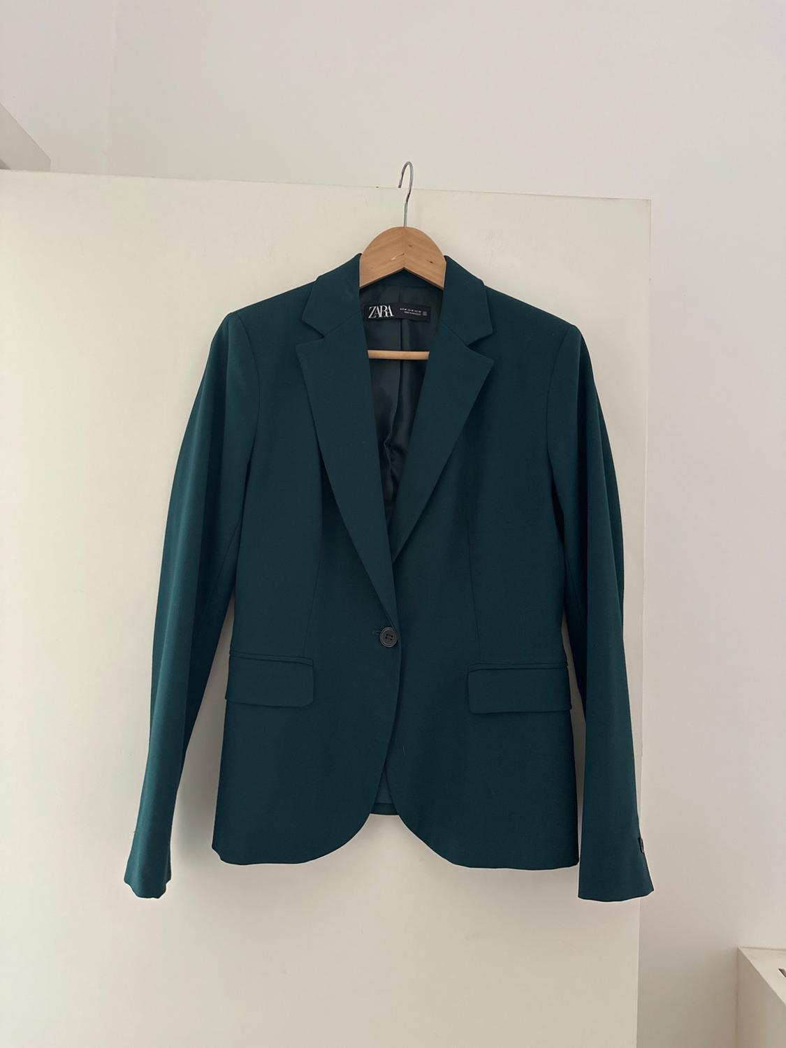 Dark green jacket hanging on a wooden hanger against a white background. Visible label: "ZARA".