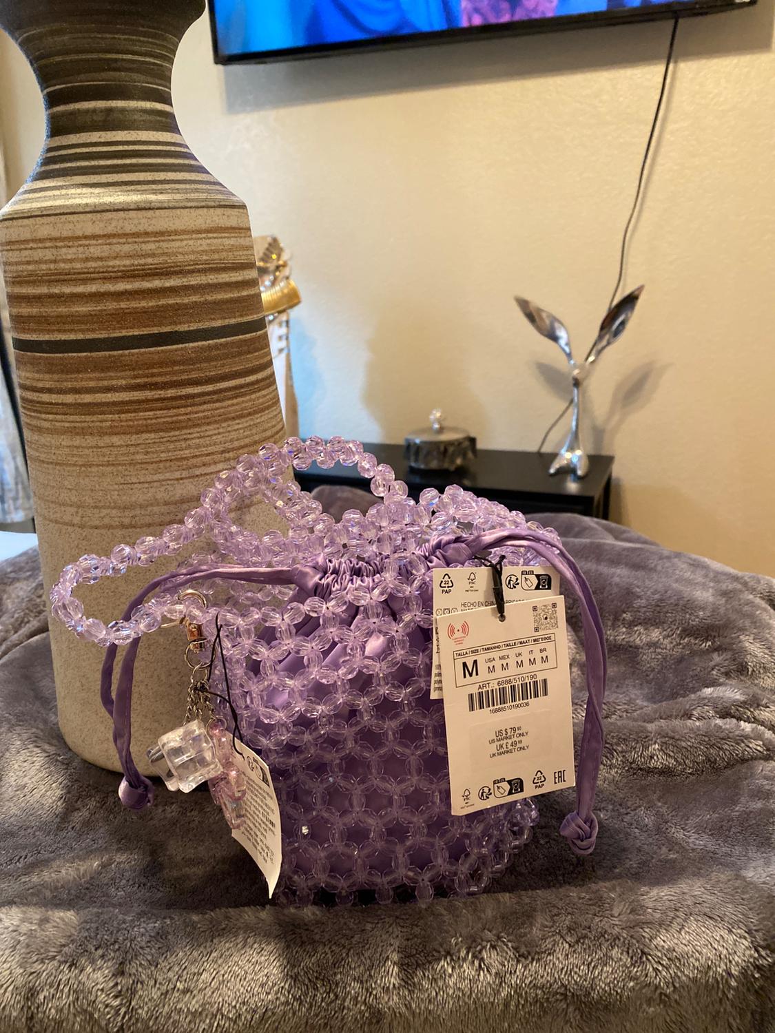 Purple beaded handbag on a grey blanket, next to a striped vase. Visible tags with price and size details. Television in the background.