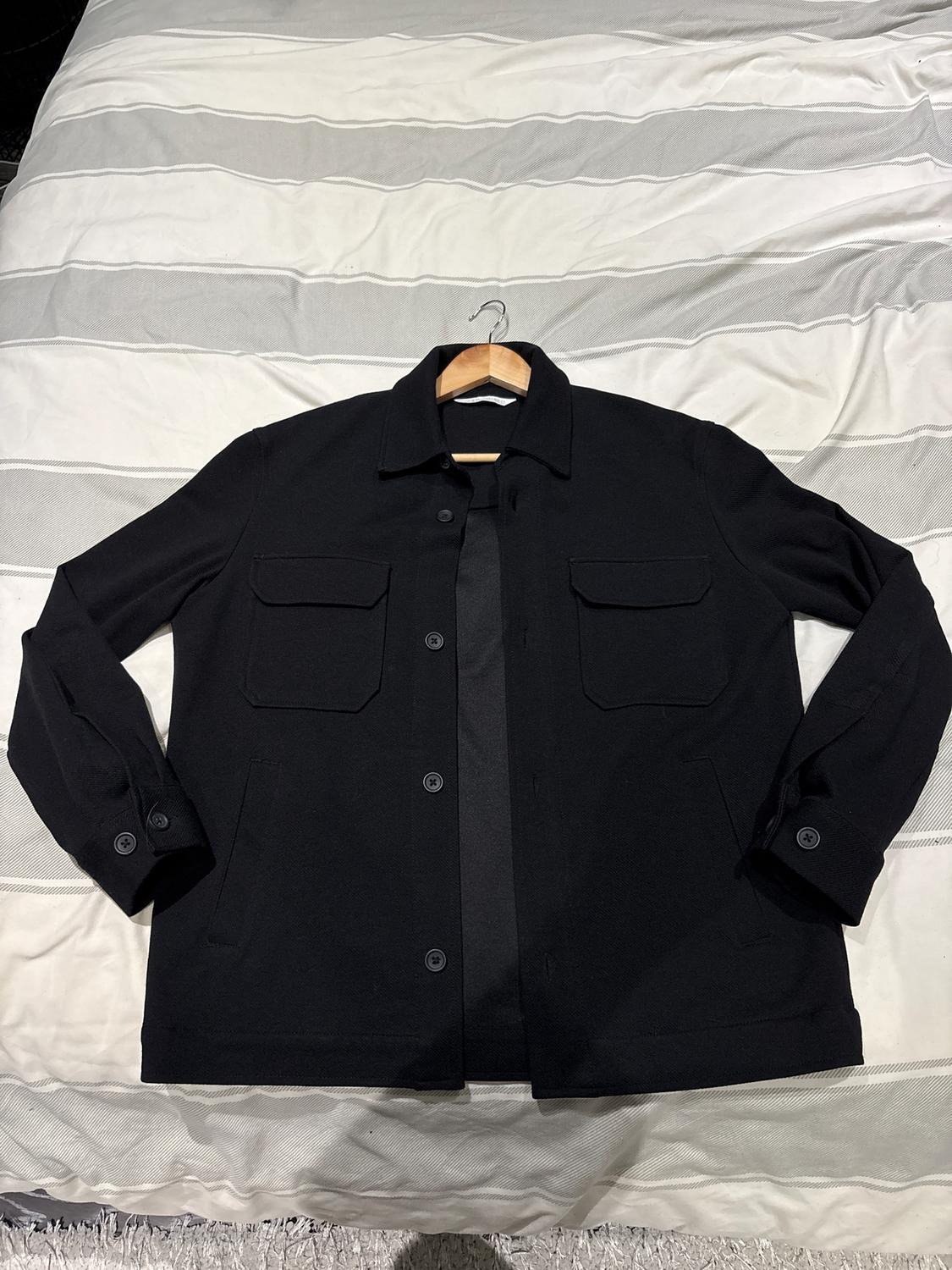 Black button-up jacket with front pockets, placed on a bed with a grey and white striped bedspread.