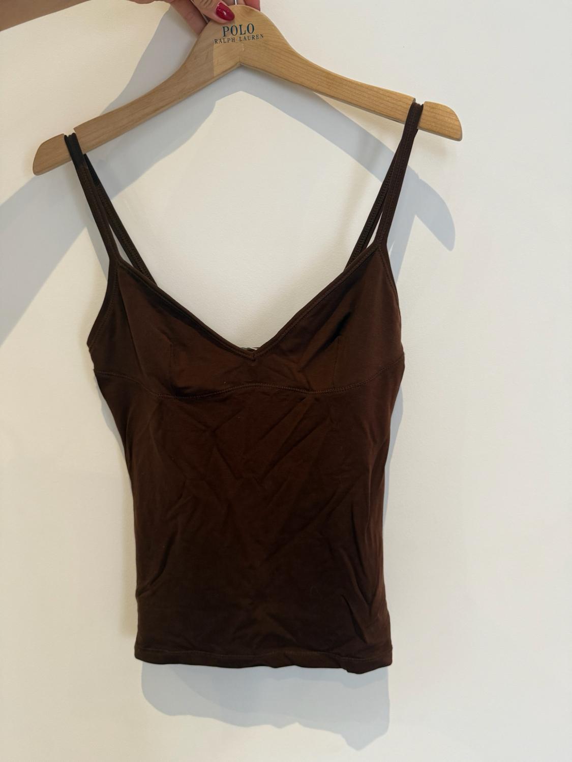 Against a white background, a hand with red nails holds a wooden hanger bearing the text POLO RALPH LAUREN, from which a brown strappy top hangs.