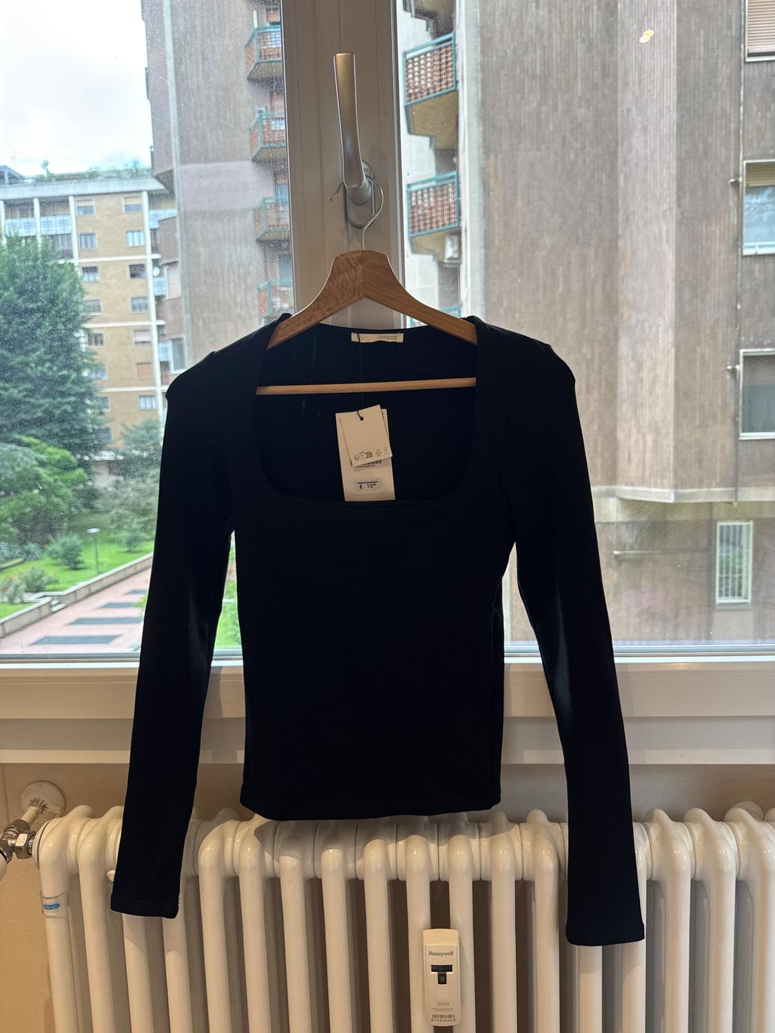 Black long-sleeved, square-neck top hanging in a window with a city view over a radiator. Text: €15, Honeywell.