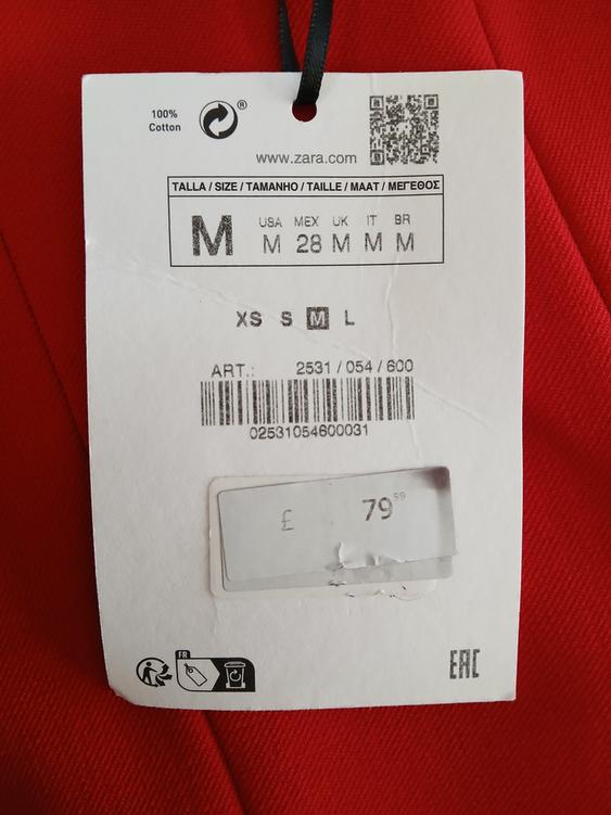 Clothing label on red fabric, shows size M, 100% cotton, price £79.99. Includes barcode and website www.zara.com.