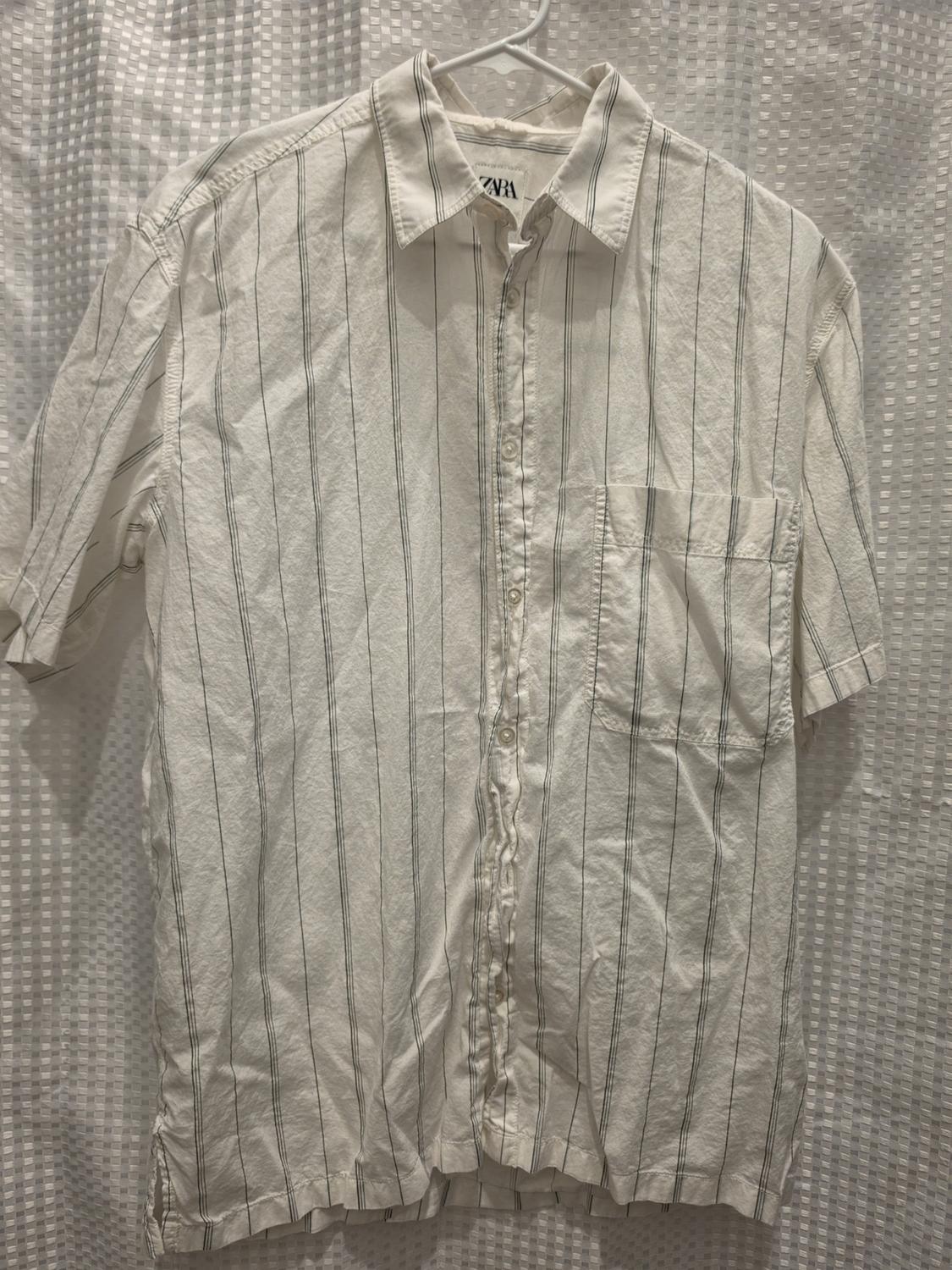 Short-sleeved white shirt with dark vertical stripes and a pocket, hanging on a hanger against a textured background. ZARA label.