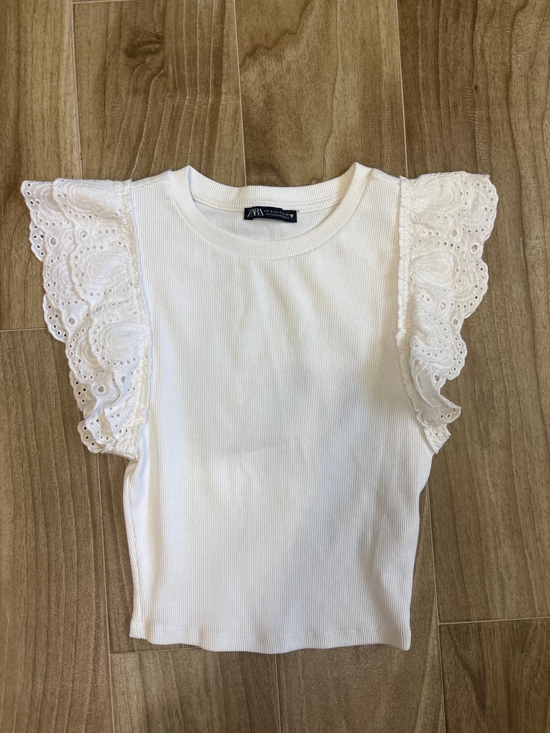 White short-sleeved blouse with lace ruffles on the sleeves, on a wooden floor. Visible tag on the collar.