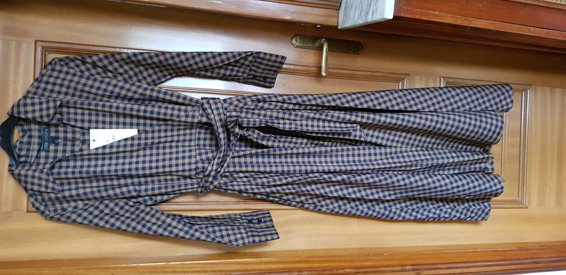Long brown and blue checked coat hanging on a wooden door, with a matching belt and a visible white label.