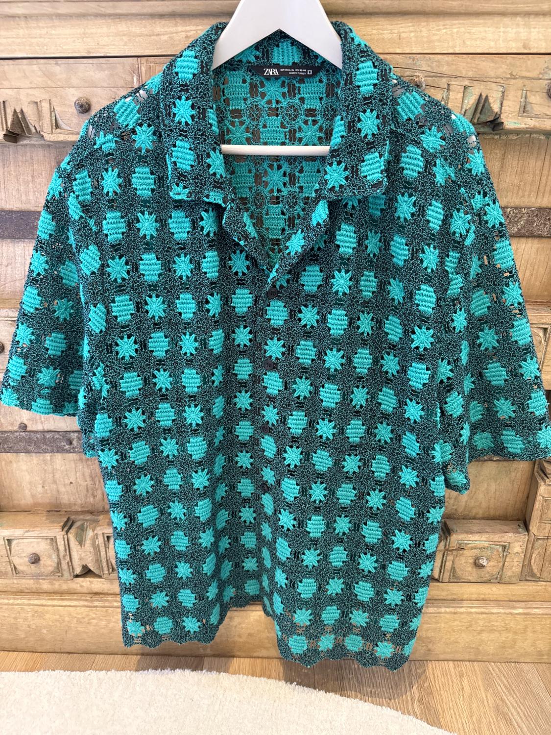 Crochet shirt in turquoise and black tones with a star pattern, hanging on a white hanger in front of a wooden background.